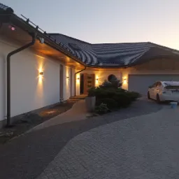 Oświetlona elewacja domu jednorodzinnego z podjazdem z kostki brukowej, widoczny fragment dachu, zaparkowany samochód i leżący pies rasy Akita Inu.