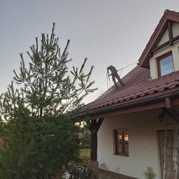 Usługi remontowo-budowlane Łukasz Lamparski - Pracownik w kombinezonie zabezpieczającym myje dach domu jednorodzinnego za pomocą myjki ciśnieniowej, widoczny fragment ogrodu i wysoka sosna.