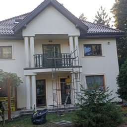 Usługi remontowo-budowlane Łukasz Lamparski - Dwupiętrowy dom z balkonem podczas renowacji elewacji, widoczne rusztowanie i pracownik w żółtej koszulce malujący nad wejściem, otoczenie domu porośnięte drzewami.