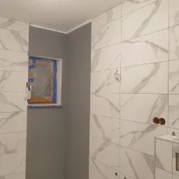 Wnętrze łazienki w trakcie remontu: białe płytki imitujące marmur na ścianach, szara ściana, okno zabezpieczone folią malarską, widoczne przygotowania do montażu armatury.