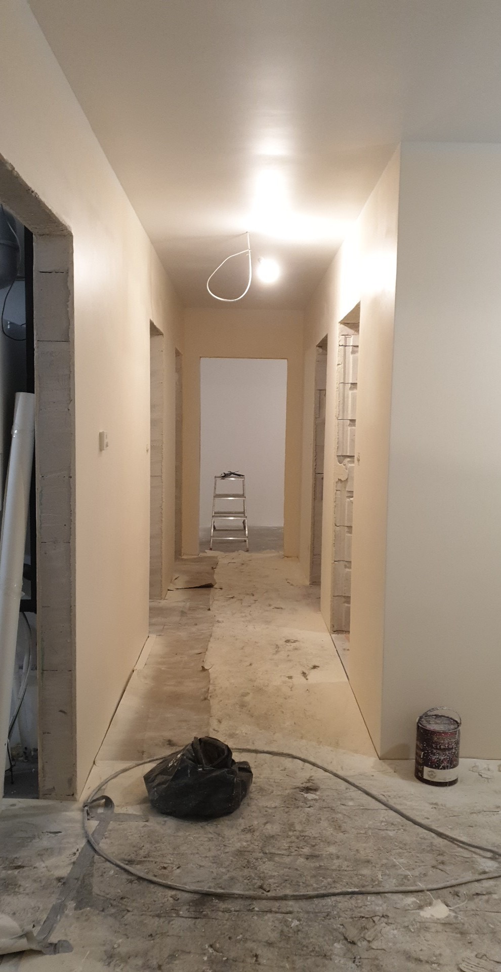 Korytarz w trakcie malowania na beżowo, widoczna drabina w głębi, puszka z farbą i worki na podłodze.