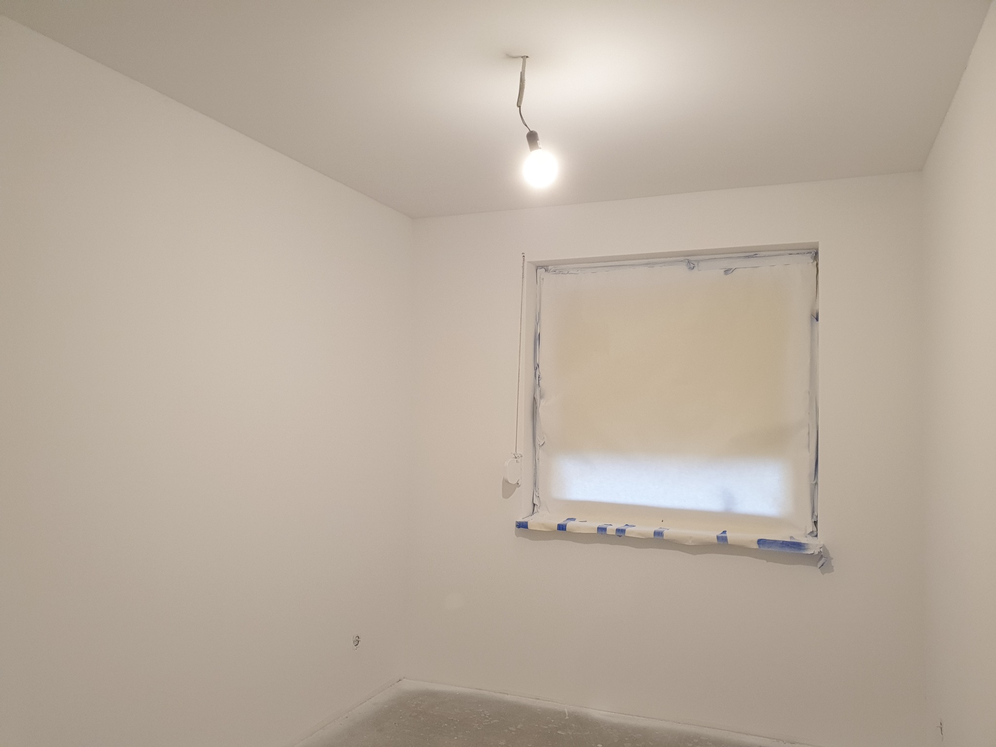 Pokój w trakcie odświeżania, ściany pomalowane na biało, okno zabezpieczone folią i taśmą malarską, surowa żarówka wisi na kablu.
