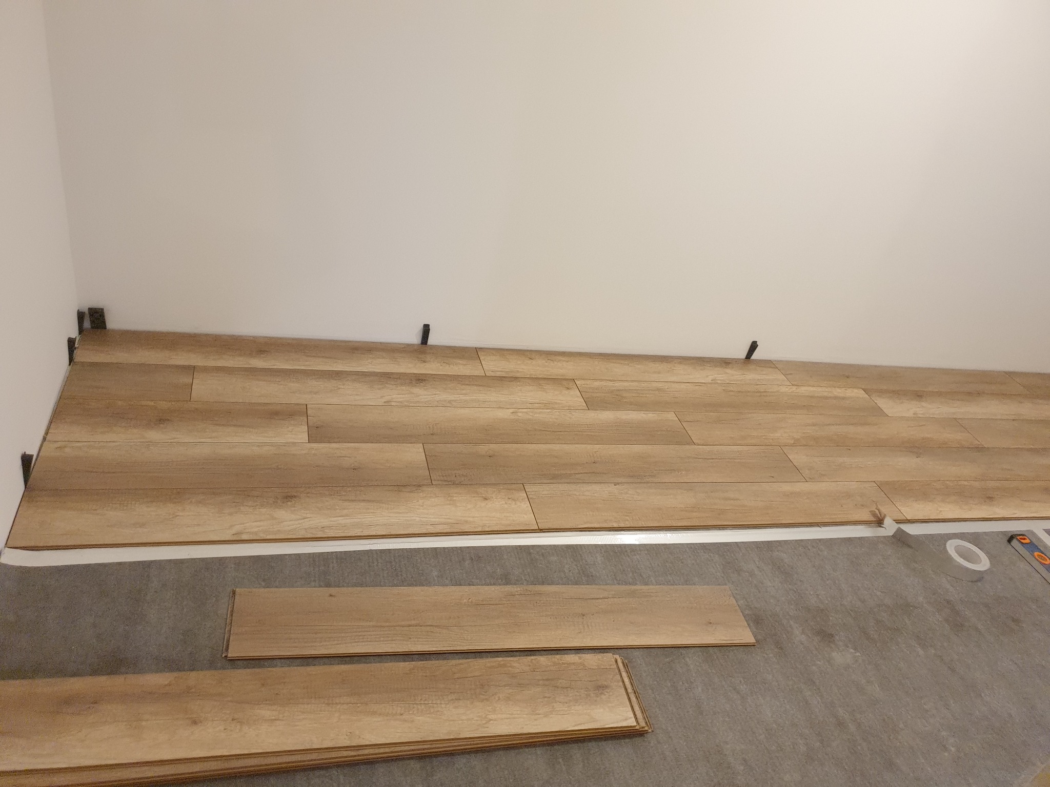 Układanie paneli podłogowych z jasnego drewna, widoczne kliny dystansowe przy ścianie, podkład oraz narzędzia pomiarowe.