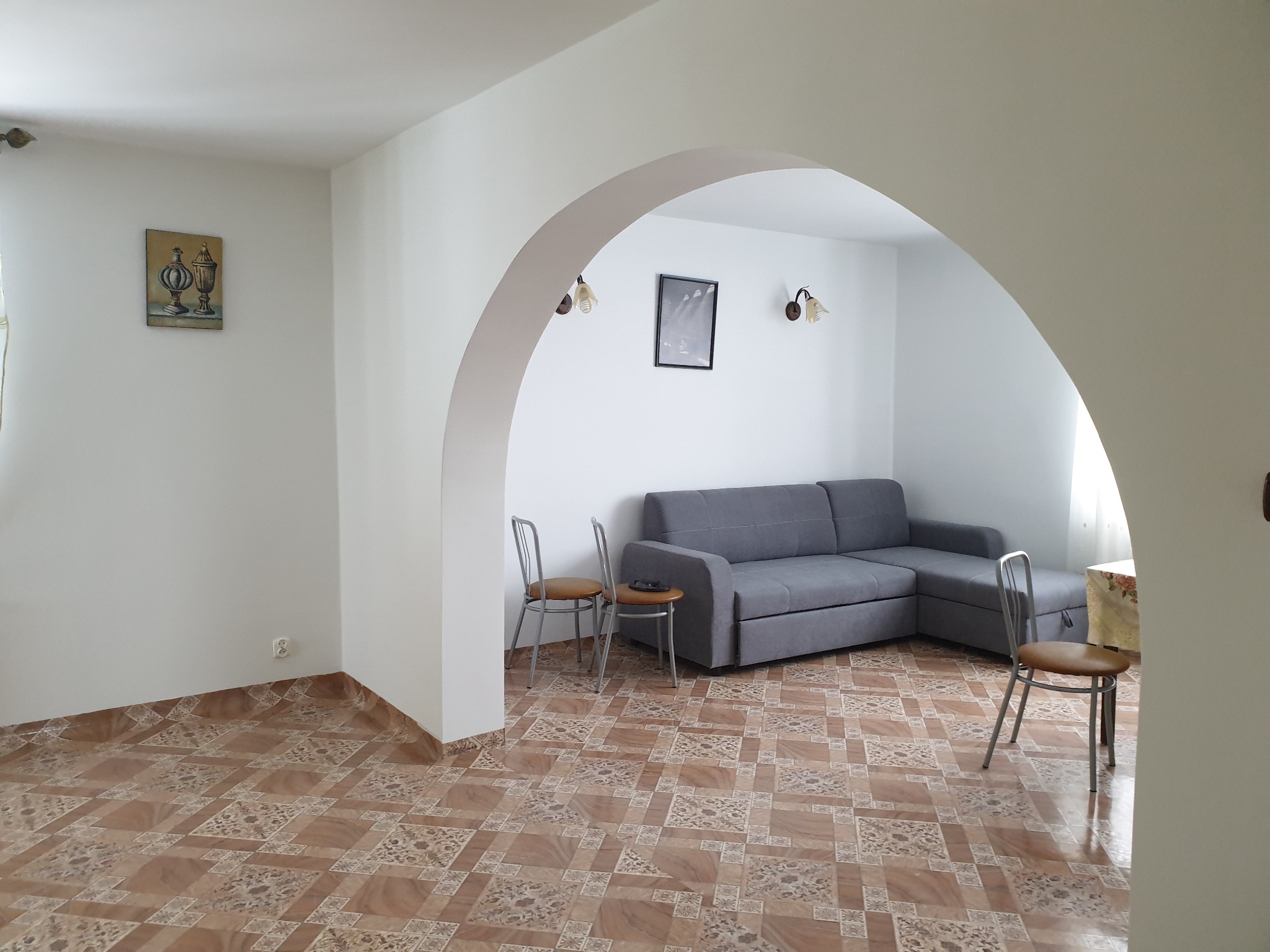 Salon z szarą narożną kanapą, dwoma krzesłami i łukowym przejściem, dekorowany obrazem i kinkietami.