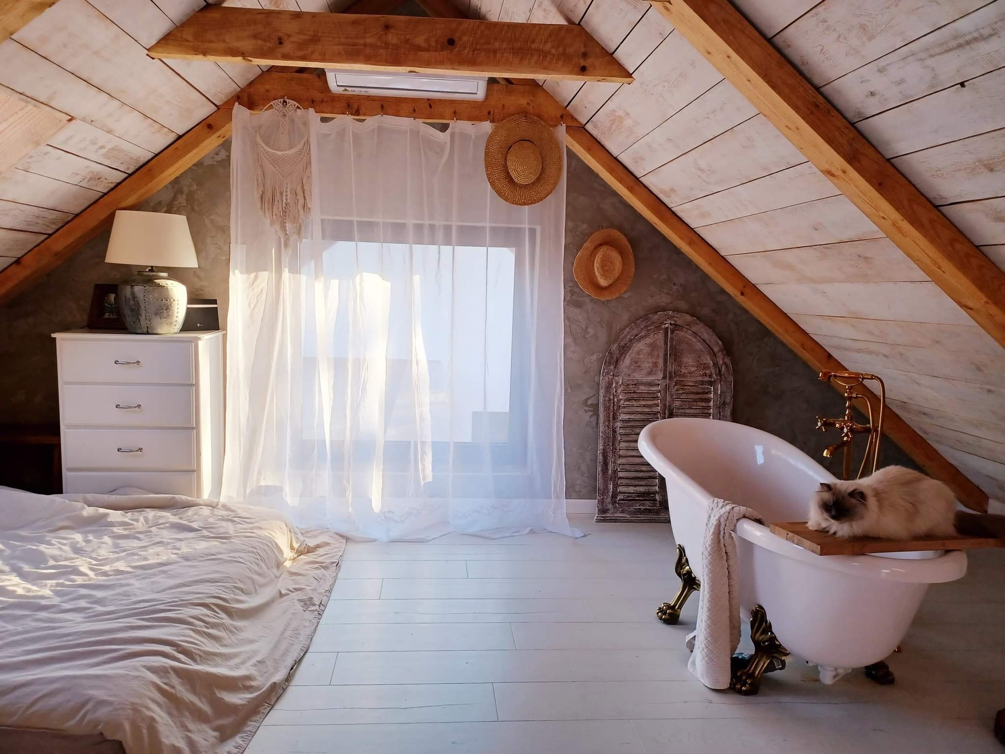 Poddasze zaadaptowane na sypialnię z wanną na złotych nóżkach, kot leży na desce ułożonej na wannie, pod skosem drewniana boazeria, na belce pod sufitem zamontowany klimatyzator.