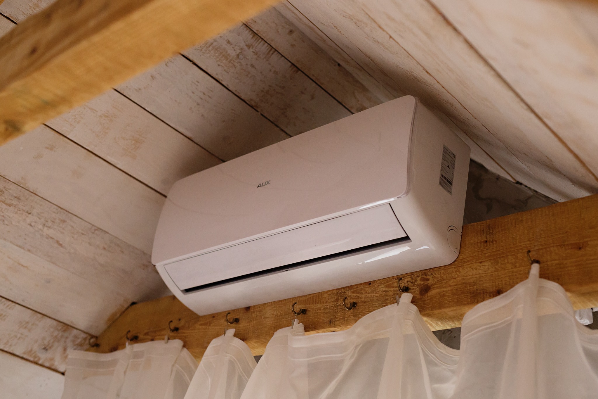 Biały klimatyzator marki AUX zamontowany pod drewnianym, skośnym sufitem z widocznymi hakami i firankami, ujęcie z dołu.