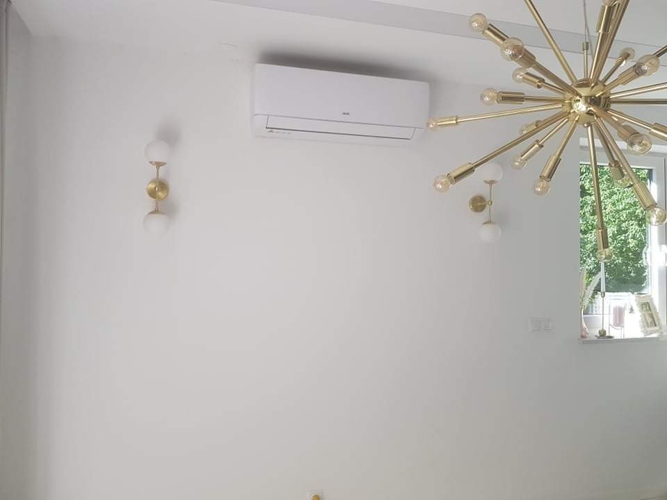 Biały klimatyzator ścienny zamontowany w jasnym pomieszczeniu z nowoczesnym złotym żyrandolem i kinkietami, widok z dołu.