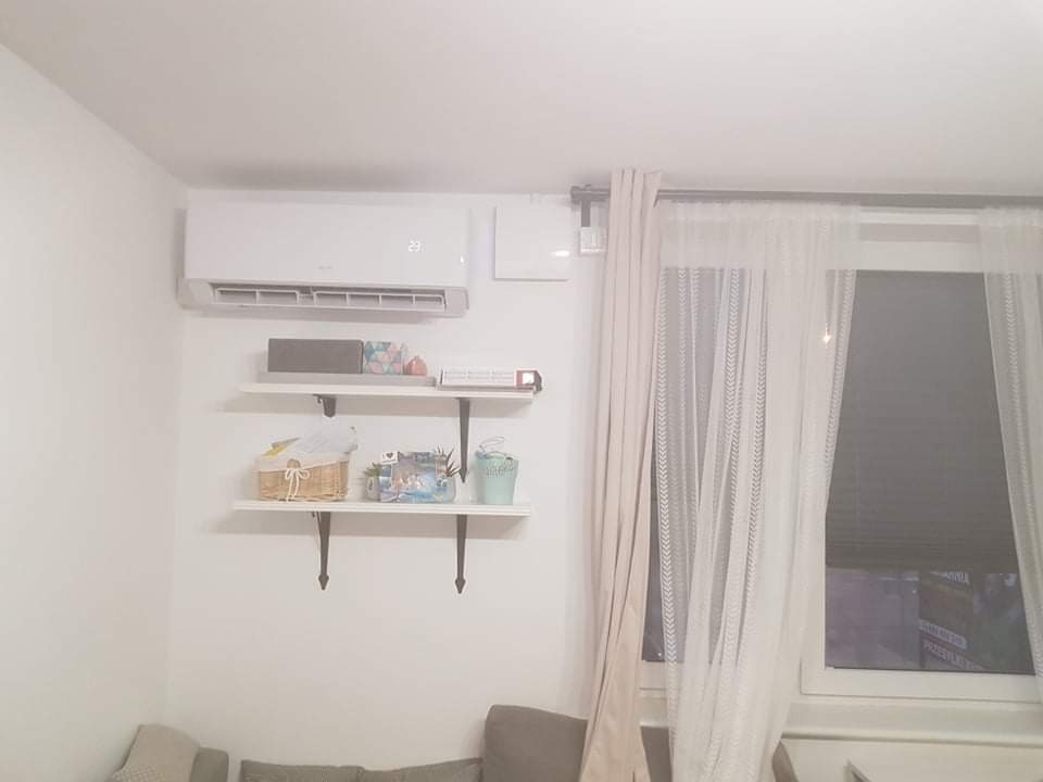 Biały klimatyzator ścienny zamontowany nad półkami z dekoracjami w jasnym pokoju z oknem zasłoniętym firankami.