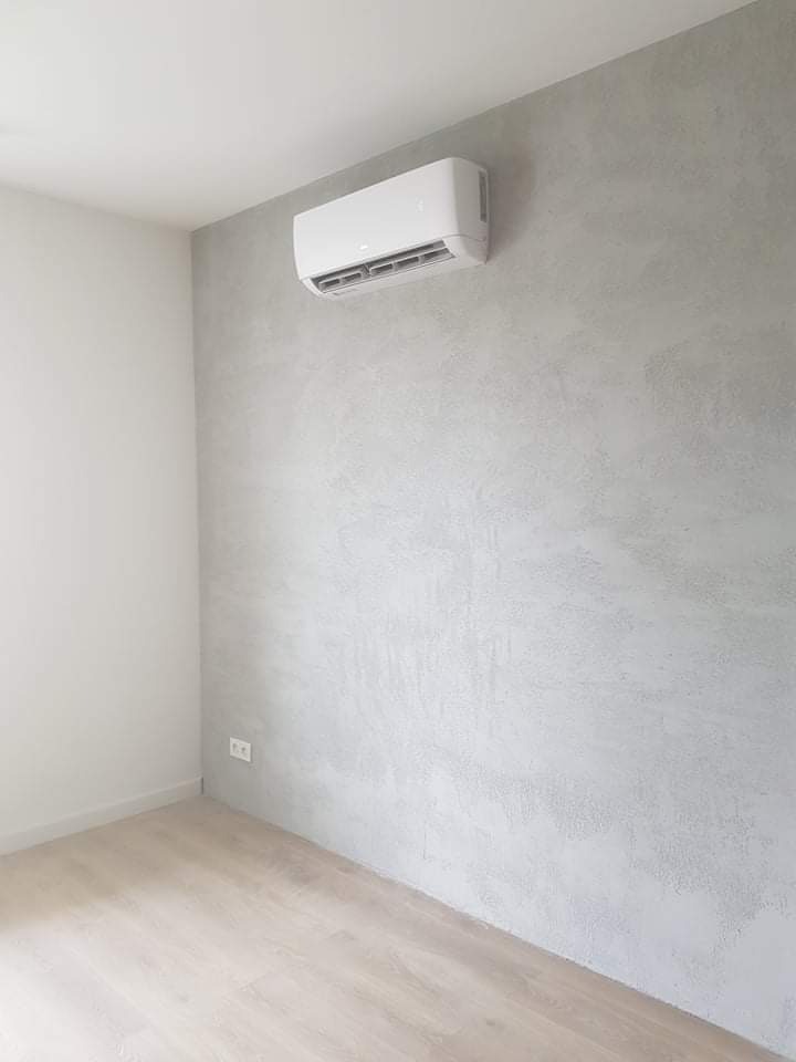 Biały klimatyzator ścienny zamontowany w rogu pokoju z szarą, teksturowaną ścianą i jasną drewnianą podłogą, widoczny gniazdko elektryczne.