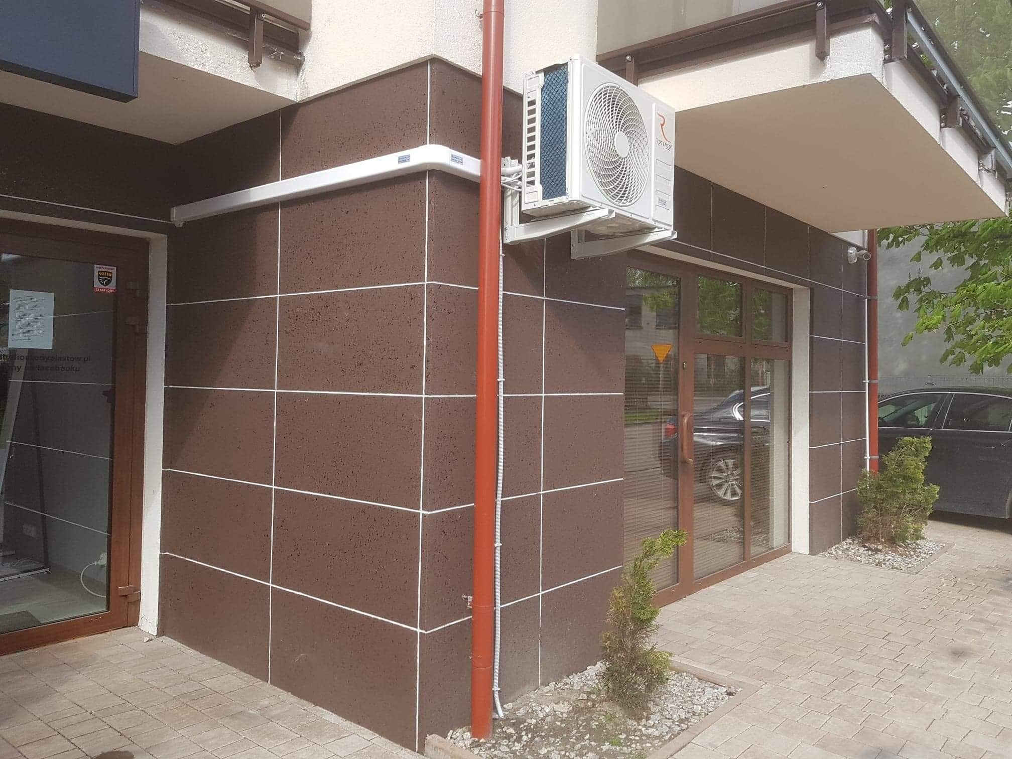 Zewnętrzna jednostka klimatyzacyjna zamontowana na brązowej ścianie budynku z widoczną instalacją elektryczną i rurami odprowadzającymi skropliny, obok okna i drzwi wejściowych.