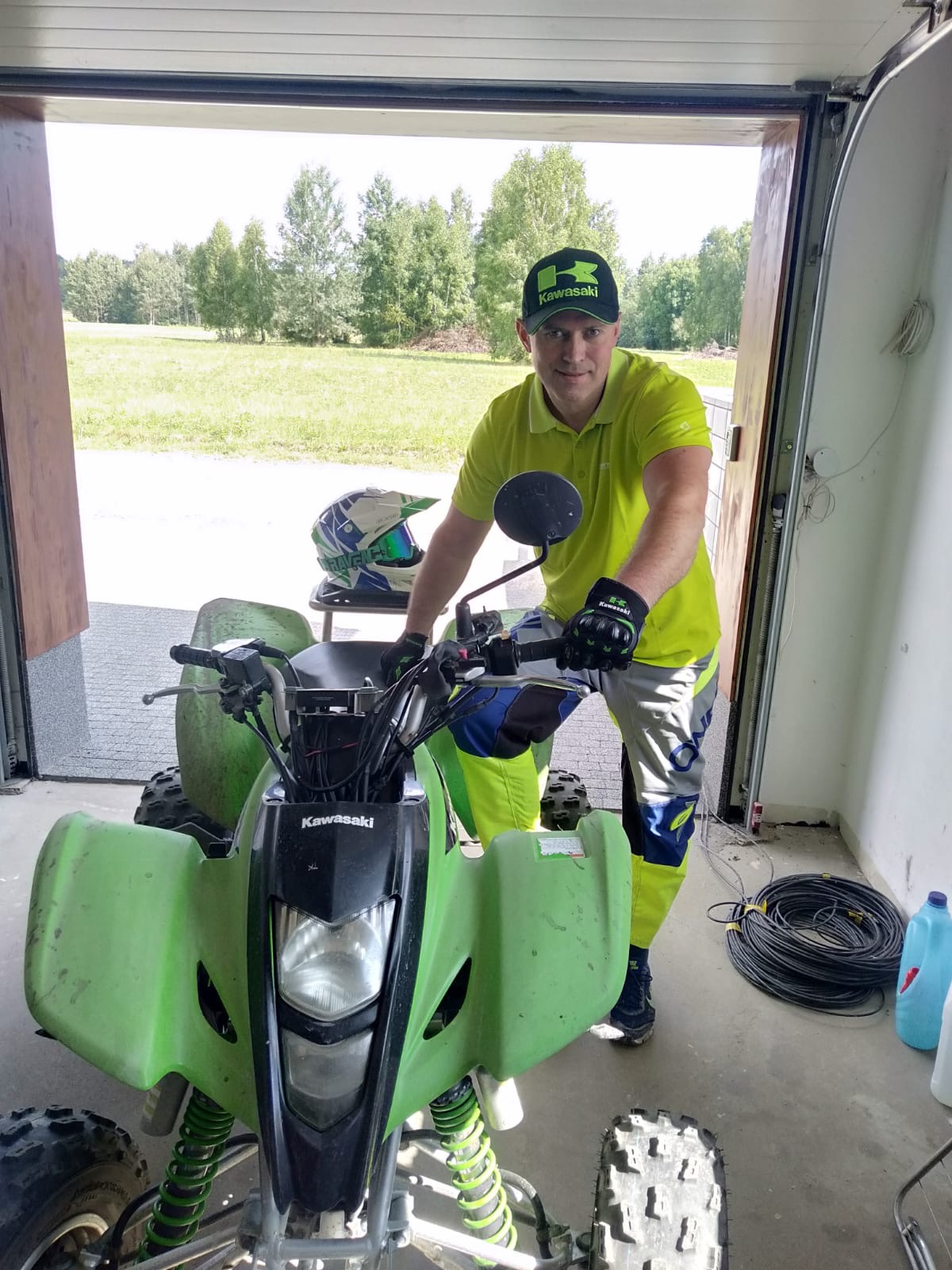 Mężczyzna w zielonej koszulce i czapce Kawasaki siedzi na zielonym quadzie Kawasaki w garażu z otwartą bramą, za którą widać drzewa i łąkę.
