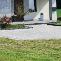 Nowo ułożona, szara kostka brukowa na podjeździe przed domem z szarą elewacją i ozdobnymi roślinami.