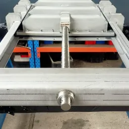 Metalowa konstrukcja ramy z profilami, widoczne spawane łączenia, śruba regulacyjna i fragment regału magazynowego w tle.