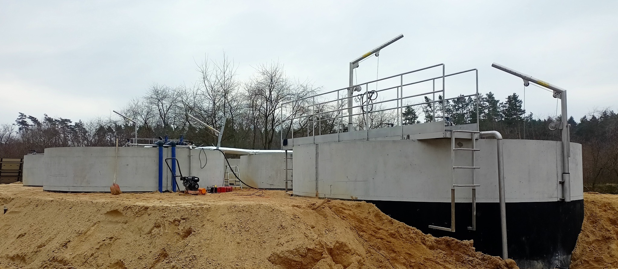 Budowa betonowych zbiorników przemysłowych na placu budowy, widoczne szalunki, drabiny i dźwigi, teren wokół zasypany piaskiem.