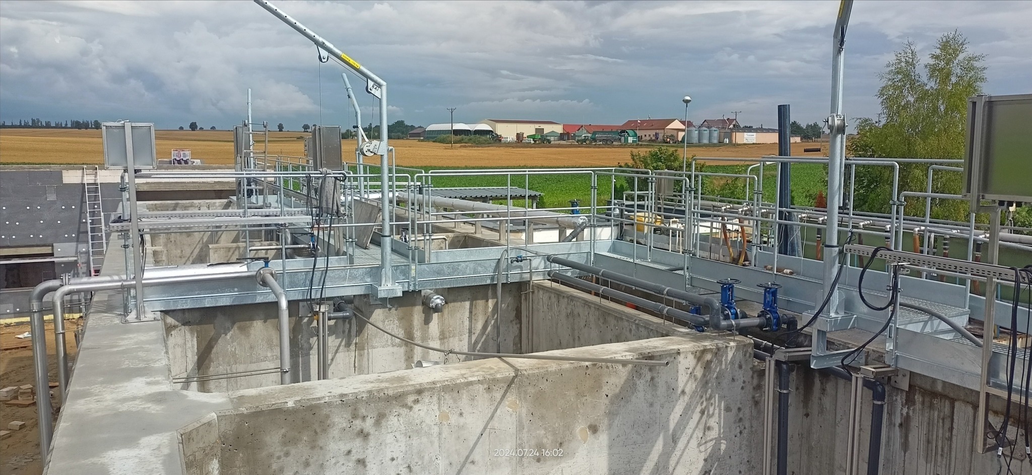 Kompleksowa instalacja przemysłowa z betonu i metalowych konstrukcji, rurociągami z zaworami, platformami dostępowymi i żurawikiem, na tle wiejskiego krajobrazu z polami i zabudowaniami.