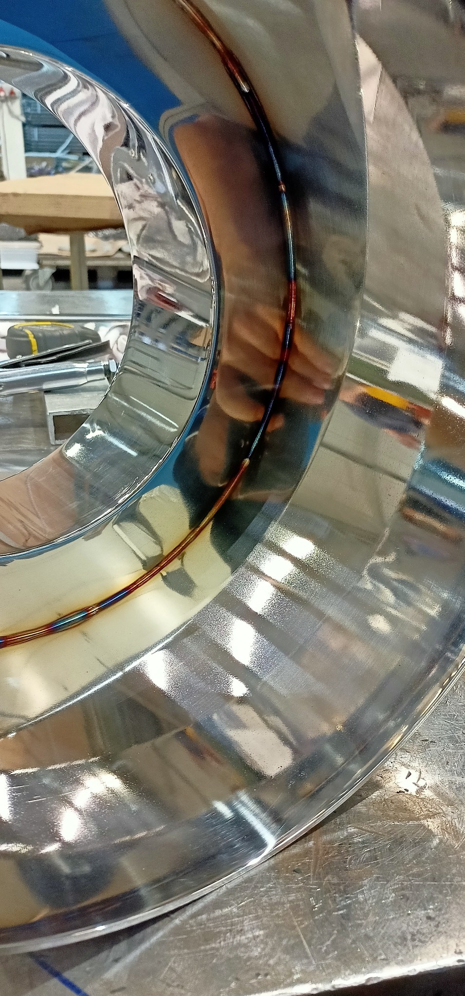 Błyszczący, spawany element z nierdzewnej stali w kształcie pierścienia, widoczne ślady spawania z charakterystycznymi przebarwieniami termicznymi, odbicie dłoni w polerowanej powierzchni.