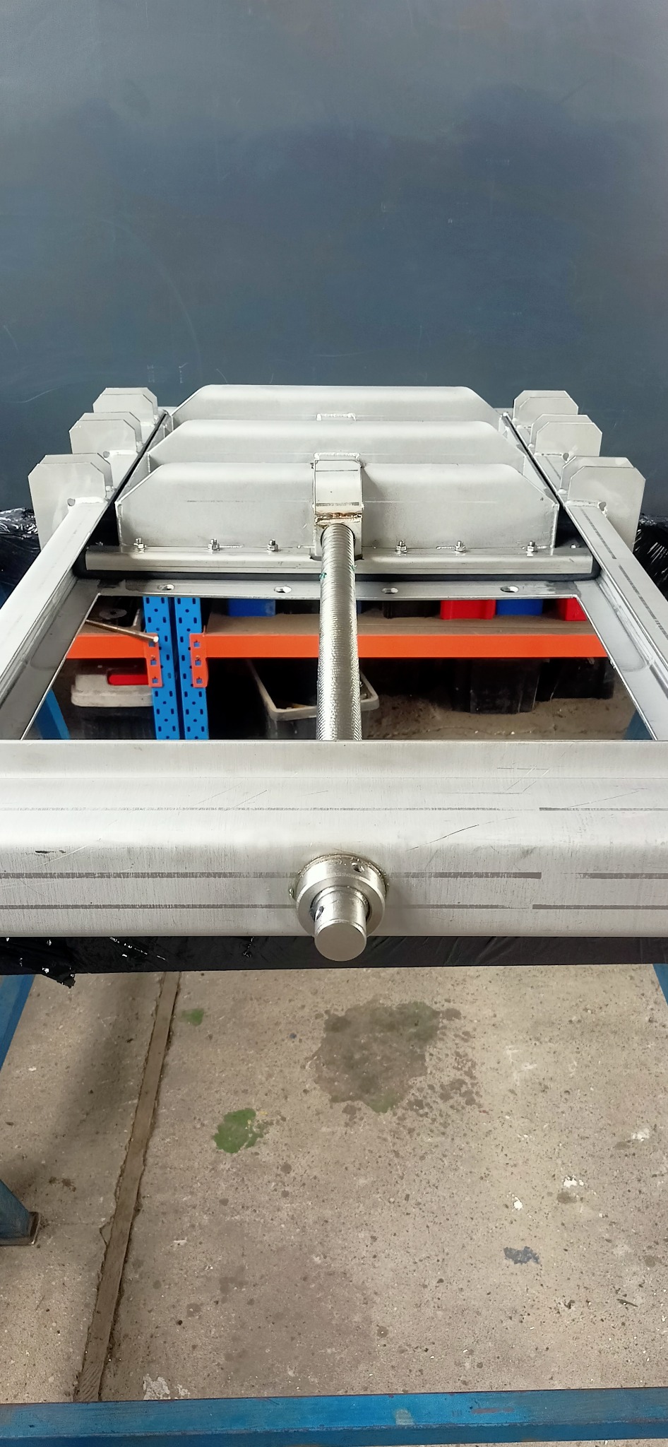 Metalowa konstrukcja ramy z profilami, widoczne spawane łączenia, śruba regulacyjna i fragment regału magazynowego w tle.