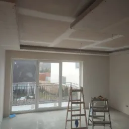 Pokój w trakcie remontu, widoczny sufit podwieszany z płyt kartonowo-gipsowych, szpachlowane łączenia, dwie drabiny, okno balkonowe z widokiem na ogród