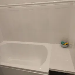 Wanna z białymi panelami ściennymi i półką, na której leży miseczka z gąbkami do mycia, minimalistyczny wystrój.