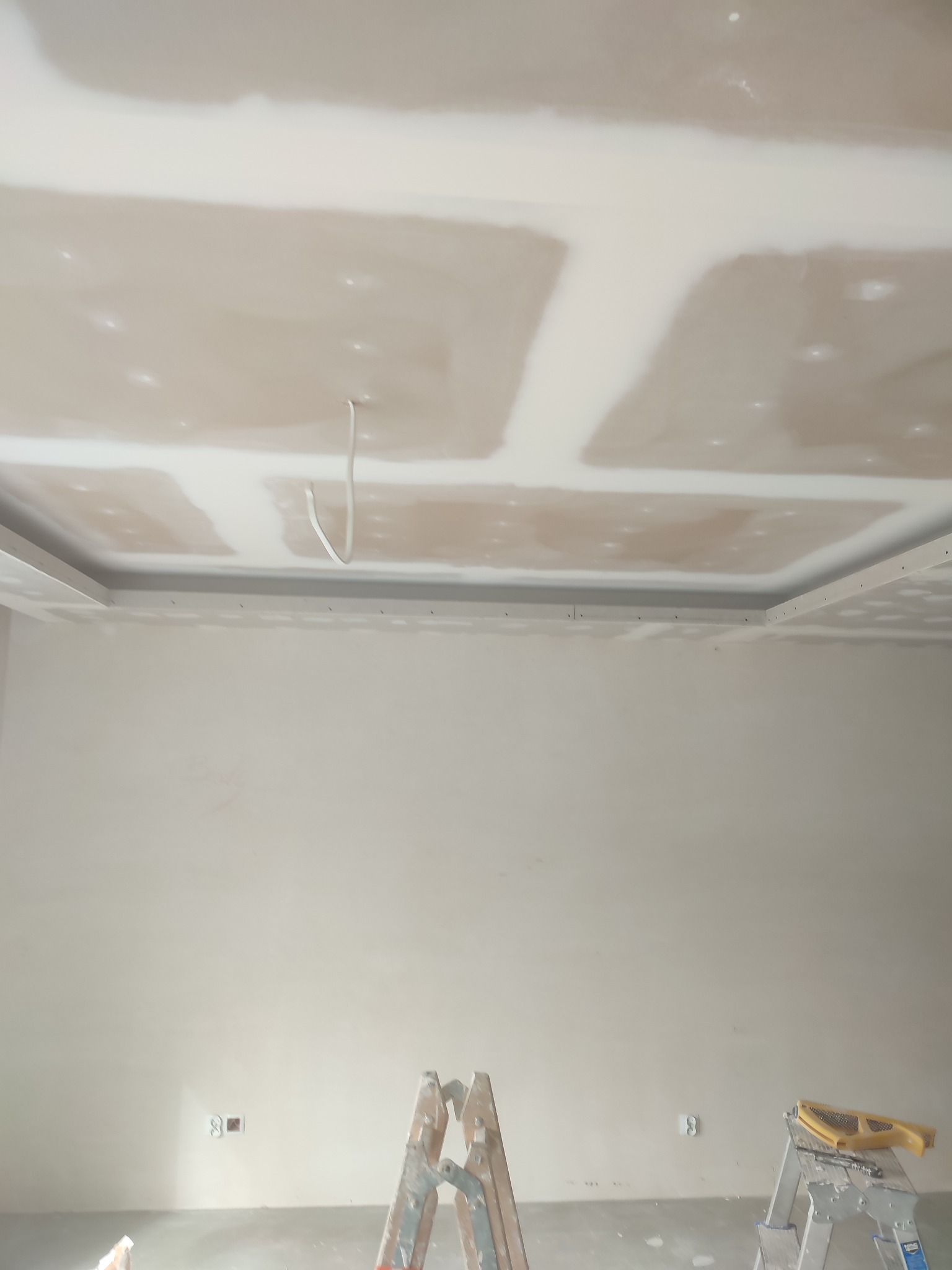 Sufit z płyt kartonowo-gipsowych w trakcie szpachlowania, widoczne łączenia płyt i wystający kabel oświetleniowy, drabina malarska i szpachelka w dolnej części kadru.