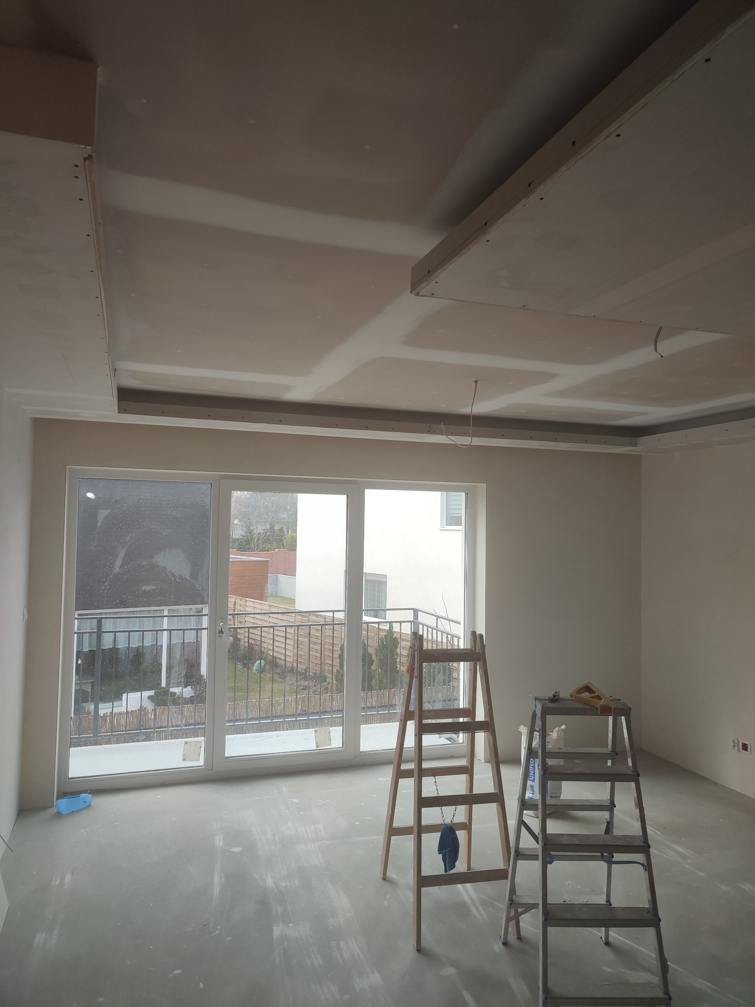 Pokój w trakcie remontu, widoczny sufit podwieszany z płyt kartonowo-gipsowych, szpachlowane łączenia, dwie drabiny, okno balkonowe z widokiem na ogród