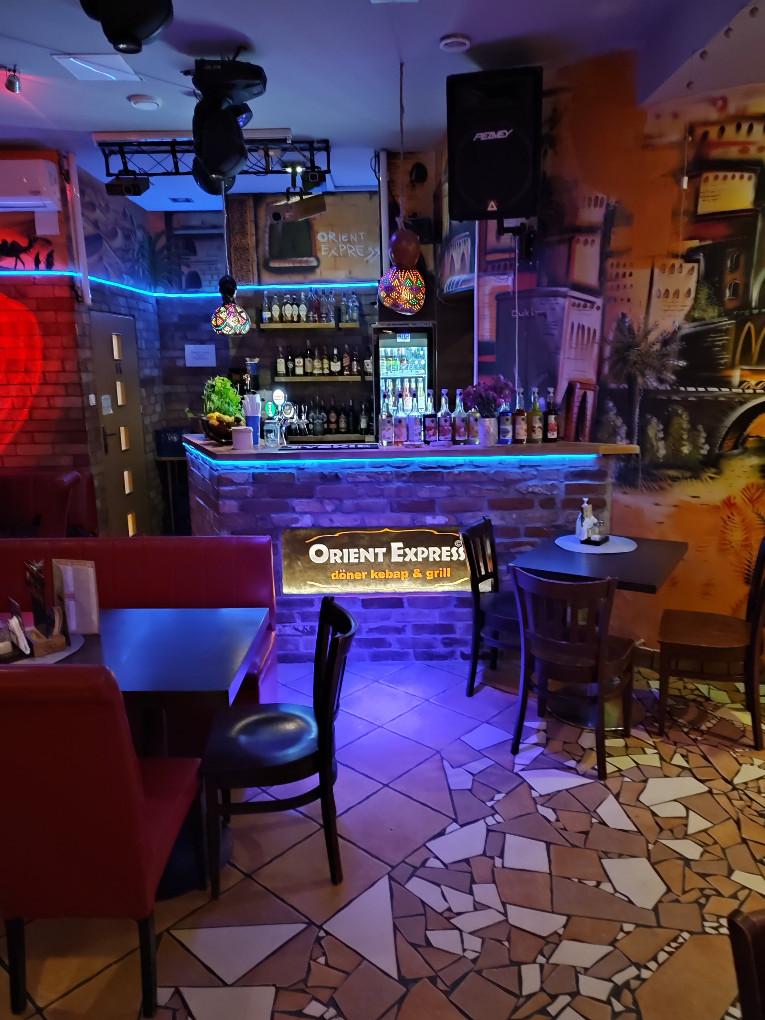 Wnętrze lokalu gastronomicznego w orientalnym stylu, z ceglanym barem podświetlonym niebieskim światłem, mozaikową podłogą i ścianą z malowidłami miejskimi.