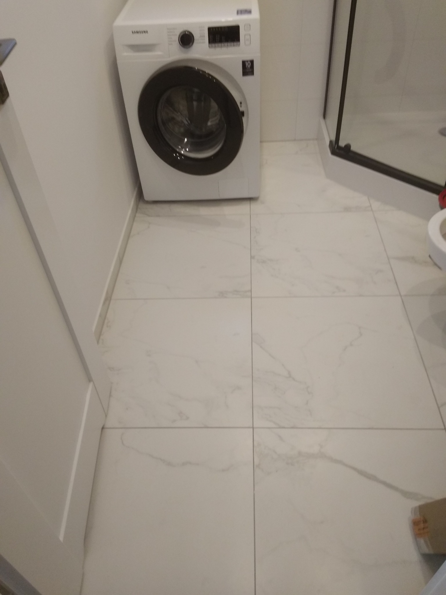 Biała pralka Samsung w narożniku łazienki z kabiną prysznicową i płytkami imitującymi marmur na podłodze.