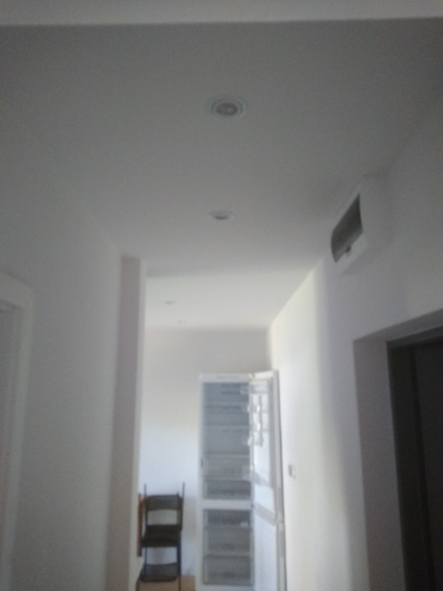 Wnętrze korytarza z pomalowanymi na biało ścianami, widoczne oświetlenie sufitowe, klimatyzator na ścianie, metalowa szafa i krzesło w tle.
