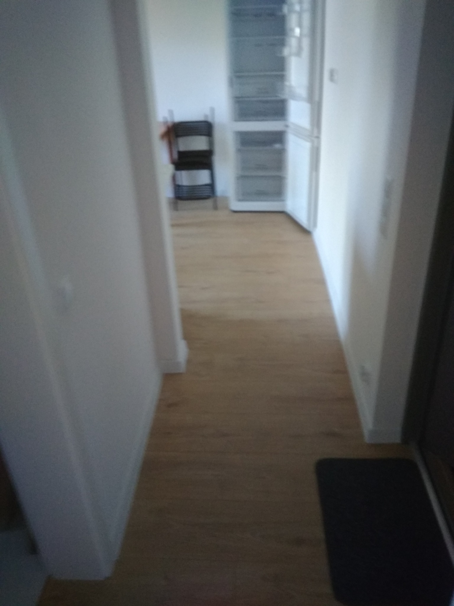 Wnętrze mieszkania z panelami podłogowymi w odcieniach brązu, widok na korytarz z białymi ścianami, w tle krzesło i lodówka.