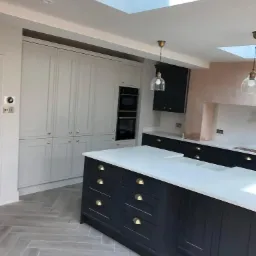 Wykończona kuchnia w stylu klasycznym z ciemnymi szafkami i białym blatem, wyspą kuchenną, jasnoszarymi szafkami w zabudowie, oświetleniem punktowym i podwieszanymi lampami oraz parkietem ułożonym...