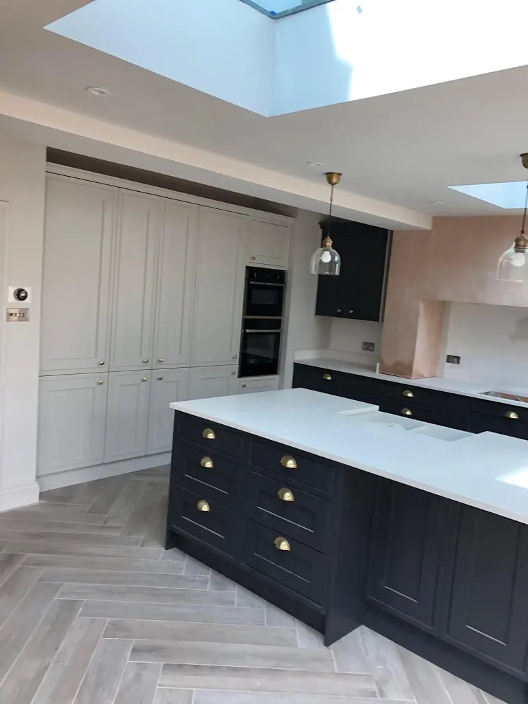 Wykończona kuchnia w stylu klasycznym z ciemnymi szafkami i białym blatem, wyspą kuchenną, jasnoszarymi szafkami w zabudowie, oświetleniem punktowym i podwieszanymi lampami oraz parkietem ułożonym...