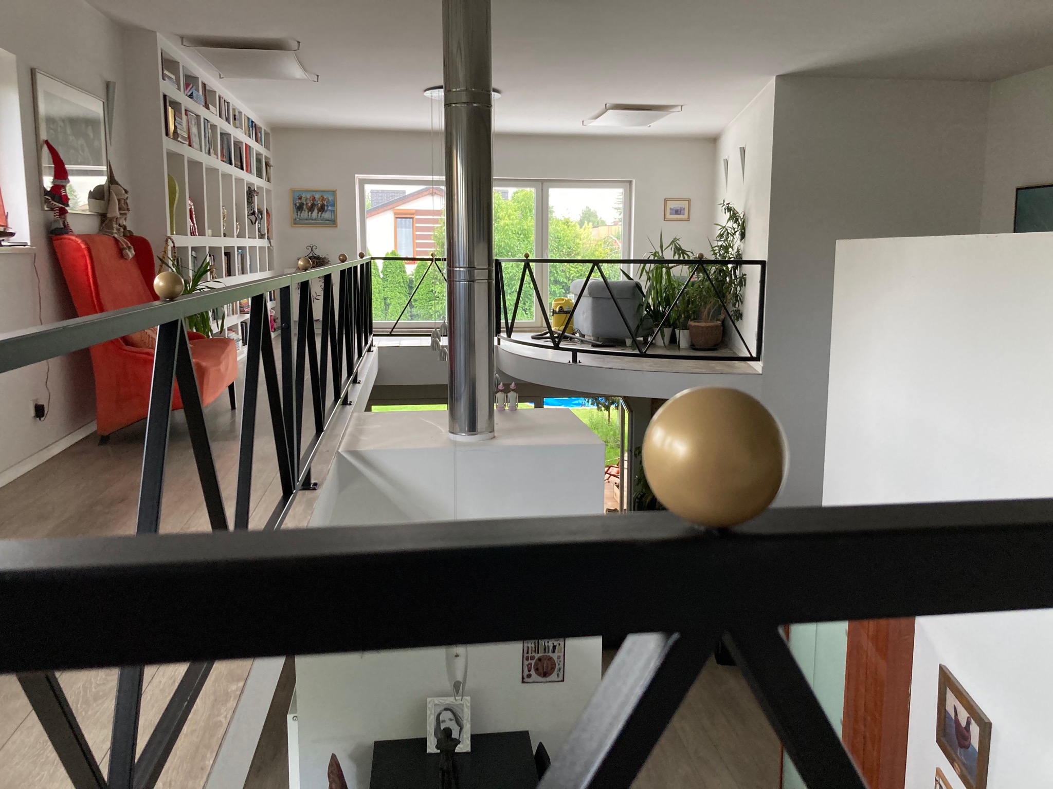 Wnętrze domu z antresolą, widok z góry przez czarną balustradę z kulkami, w tle salon z biblioteczką i okna.