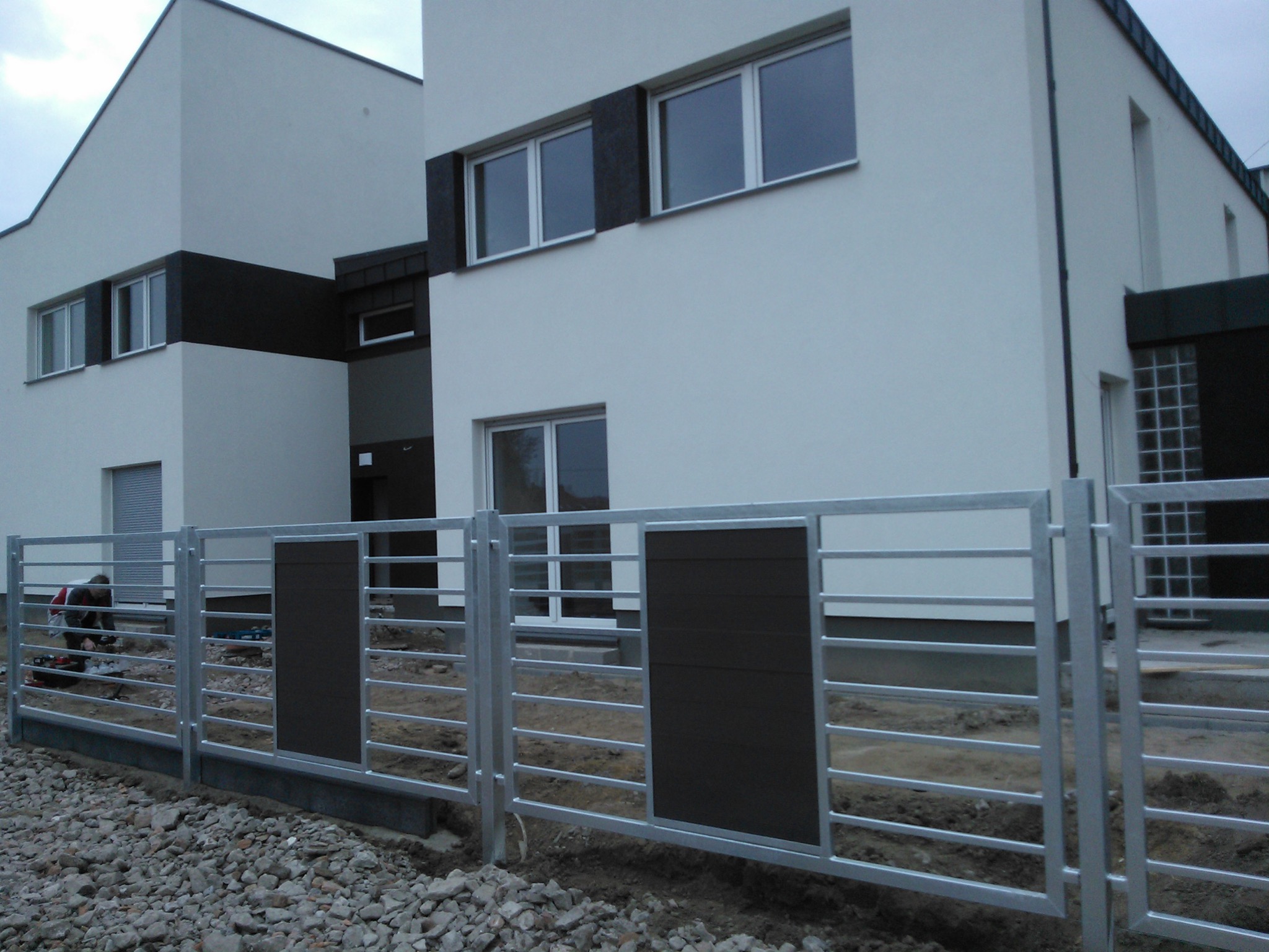 Montaż metalowego ogrodzenia panelowego z elementami drewnopodobnymi przed nowoczesnym budynkiem mieszkalnym, widoczny pracownik z narzędziami.