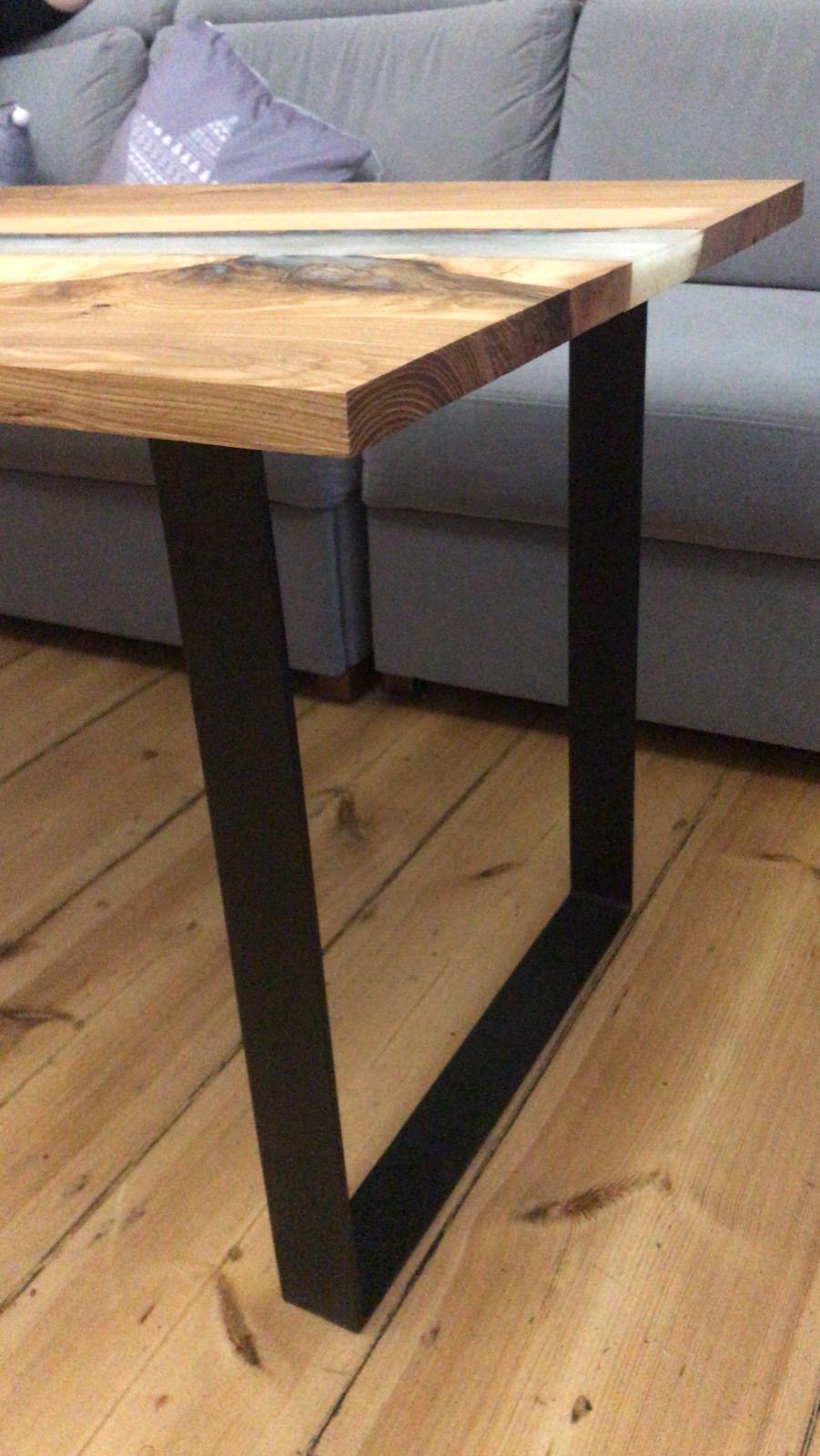 Stolik kawowy z blatem z drewna i żywicy epoksydowej, z czarnymi, metalowymi nogami w kształcie litery V, na tle szarej sofy.