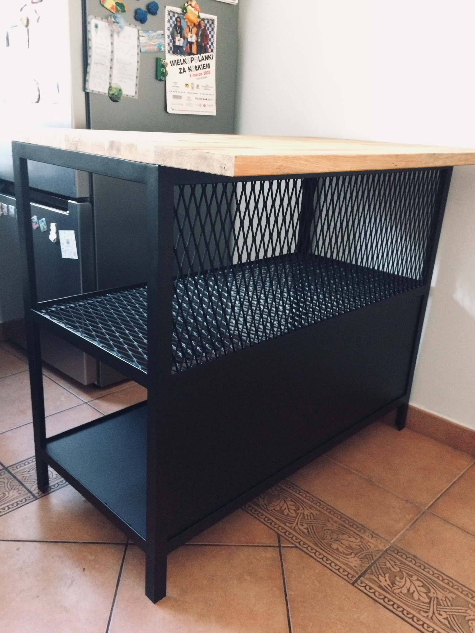 Stół kuchenny na czarnym metalowym stelażu z drewnianym blatem, widoczne metalowe siatki jako elementy konstrukcyjne, na podłodze brązowe płytki.