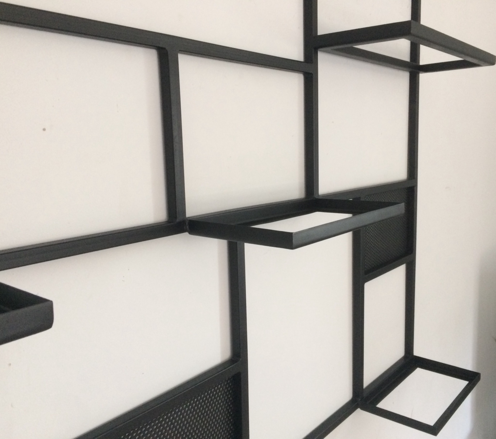 Metalowa, modułowa półka ścienna w kolorze czarnym, z elementami siatki, zamontowana na białej ścianie.