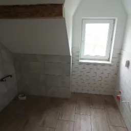 Ukończona łazienka na poddaszu ze skośnym sufitem, wykończona płytkami w dwóch odcieniach szarości, z drewnianą belką stropową i oknem dachowym.