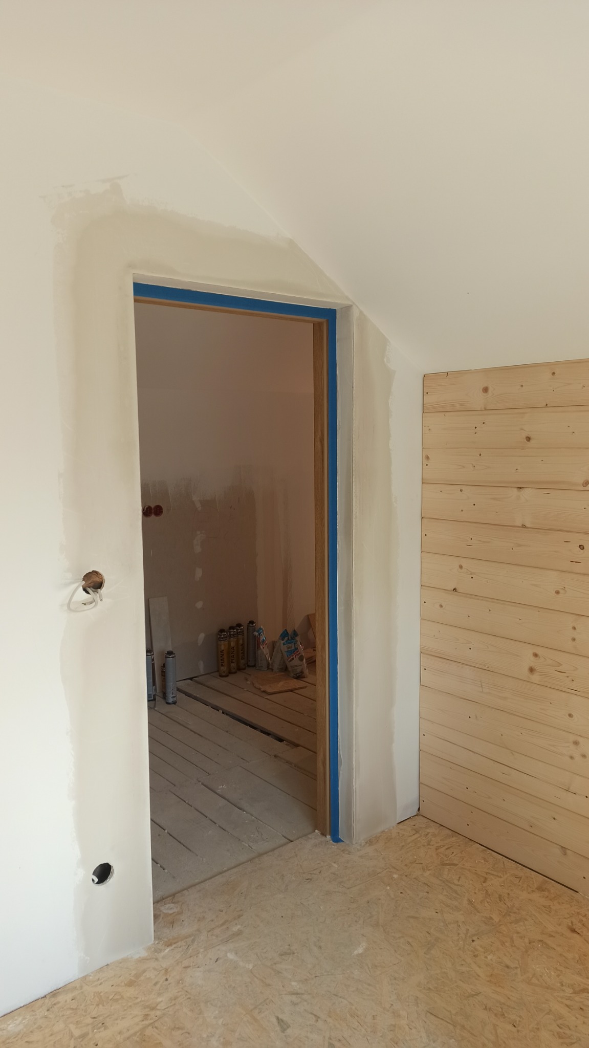 Wnętrze w trakcie remontu: ściany z świeżo położoną gładzią, zabezpieczone taśmą malarską ościeżnice drzwi, częściowo wykończona drewniana boazeria, widoczne puszki z farbą i worki z zaprawą...