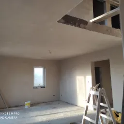 Wnętrze w trakcie prac wykończeniowych: ściany i sufit pokryte świeżo nałożoną gładzią, widoczna drabina, wiadro z farbą i niedokończone przejście w suficie.
