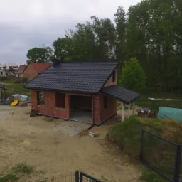 Niedokończony budynek z czerwonej cegły z czarnym dachem, widok z góry na tle drzew i innych budynków.