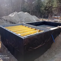 Wykonana izolacja pionowa fundamentów z widocznym deskowaniem i żółtymi belkami, wykop wokół fundamentów zasypany piaskiem, w tle las i pryzma piasku.