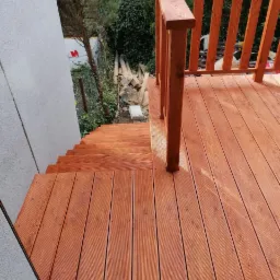 Drewniany taras z balustradą i schodami, widok z góry, deski ryflowane, świeżo po impregnacji, w tle ogród z gruzem budowlanym.