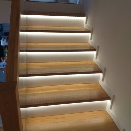 Woodcraft - Drewniane schody z podświetleniem LED, widok z dołu, jasne drewno, białe boki, nowoczesny design.