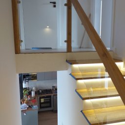 Woodcraft - Drewniane schody z podświetleniem LED i szklaną balustradą, widok z góry na fragment kuchni.