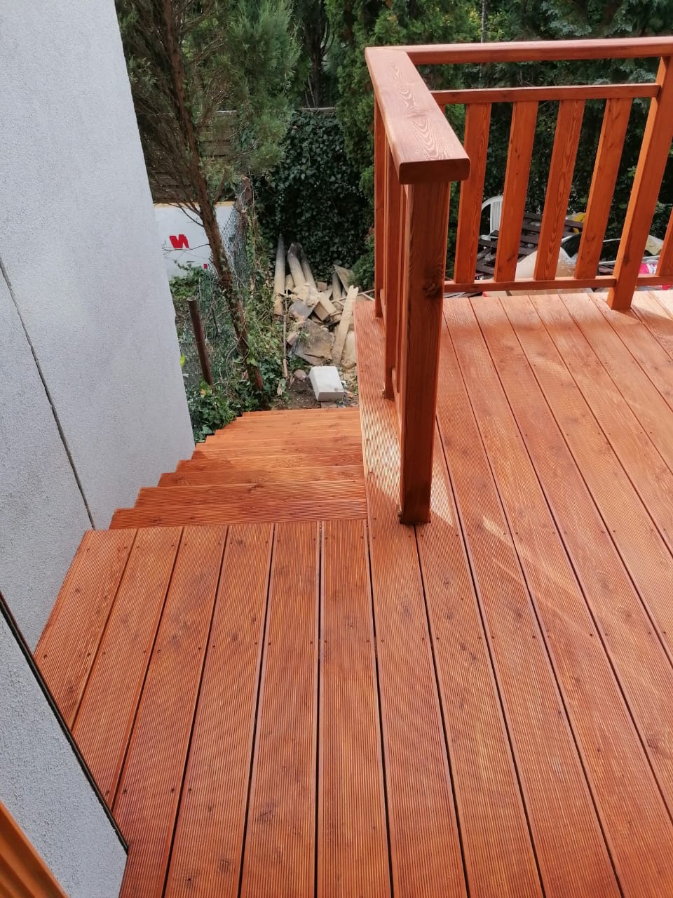 Drewniany taras z balustradą i schodami, widok z góry, deski ryflowane, świeżo po impregnacji, w tle ogród z gruzem budowlanym.