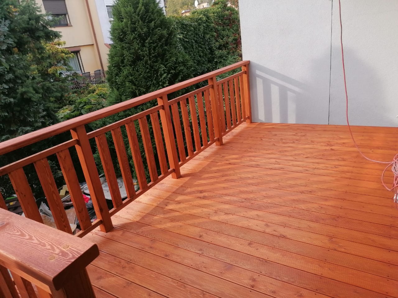 Drewniany taras z balustradą w odcieniach brązu, świeżo po impregnacji, widok z góry, w tle zieleń drzew i fragment budynku.