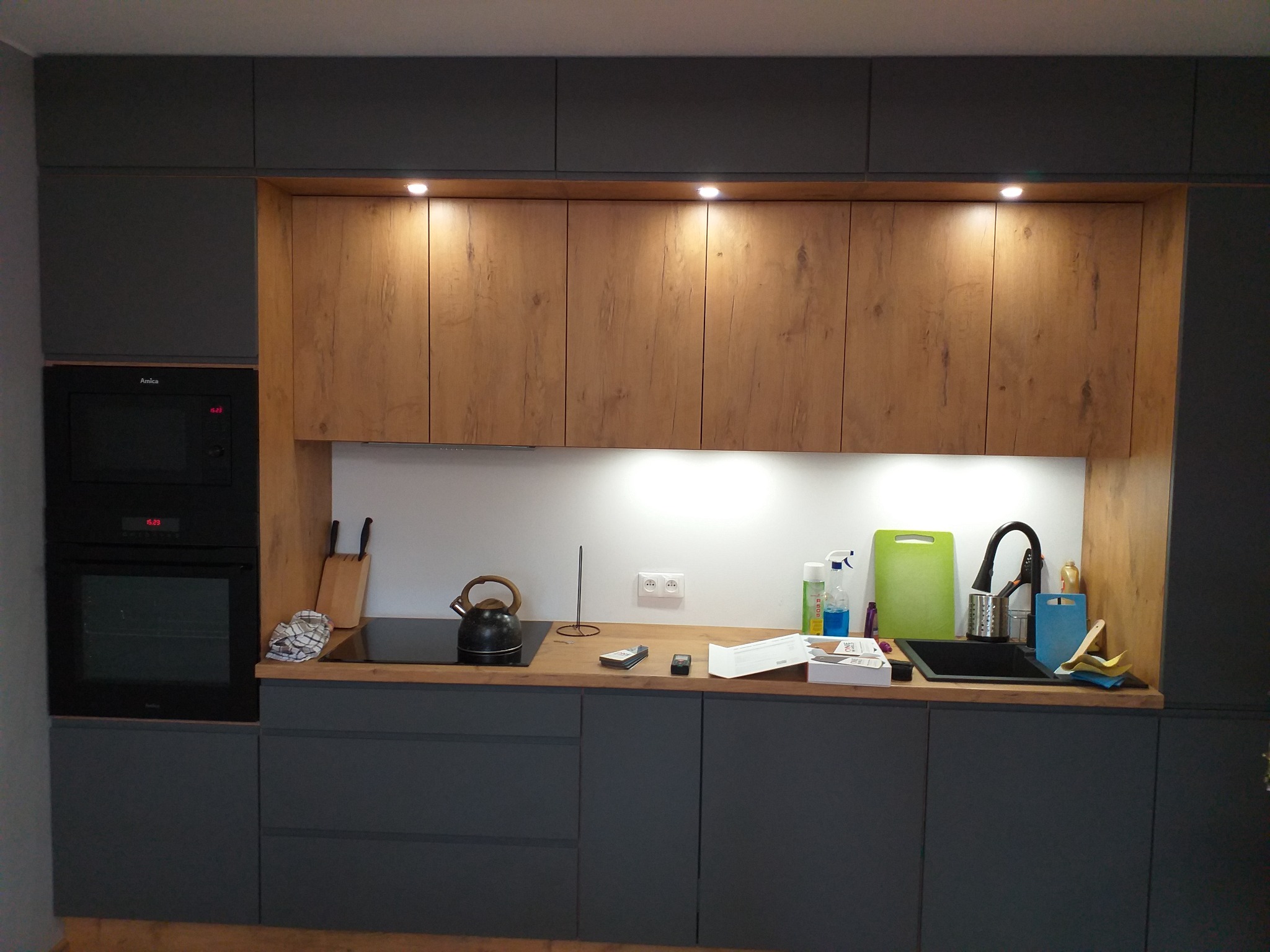 Nowoczesna kuchnia z szarymi szafkami dolnymi i drewnopodobnymi górnymi, oświetlona punktowo, z czarnym piekarnikiem i płytą grzewczą.