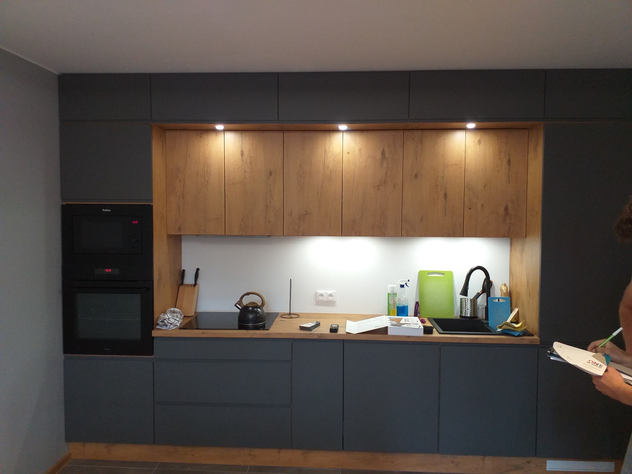 Nowoczesna kuchnia z szarymi szafkami dolnymi i drewnopodobnymi szafkami górnymi, czarny piekarnik w zabudowie, osoba z notatnikiem w kadrze.