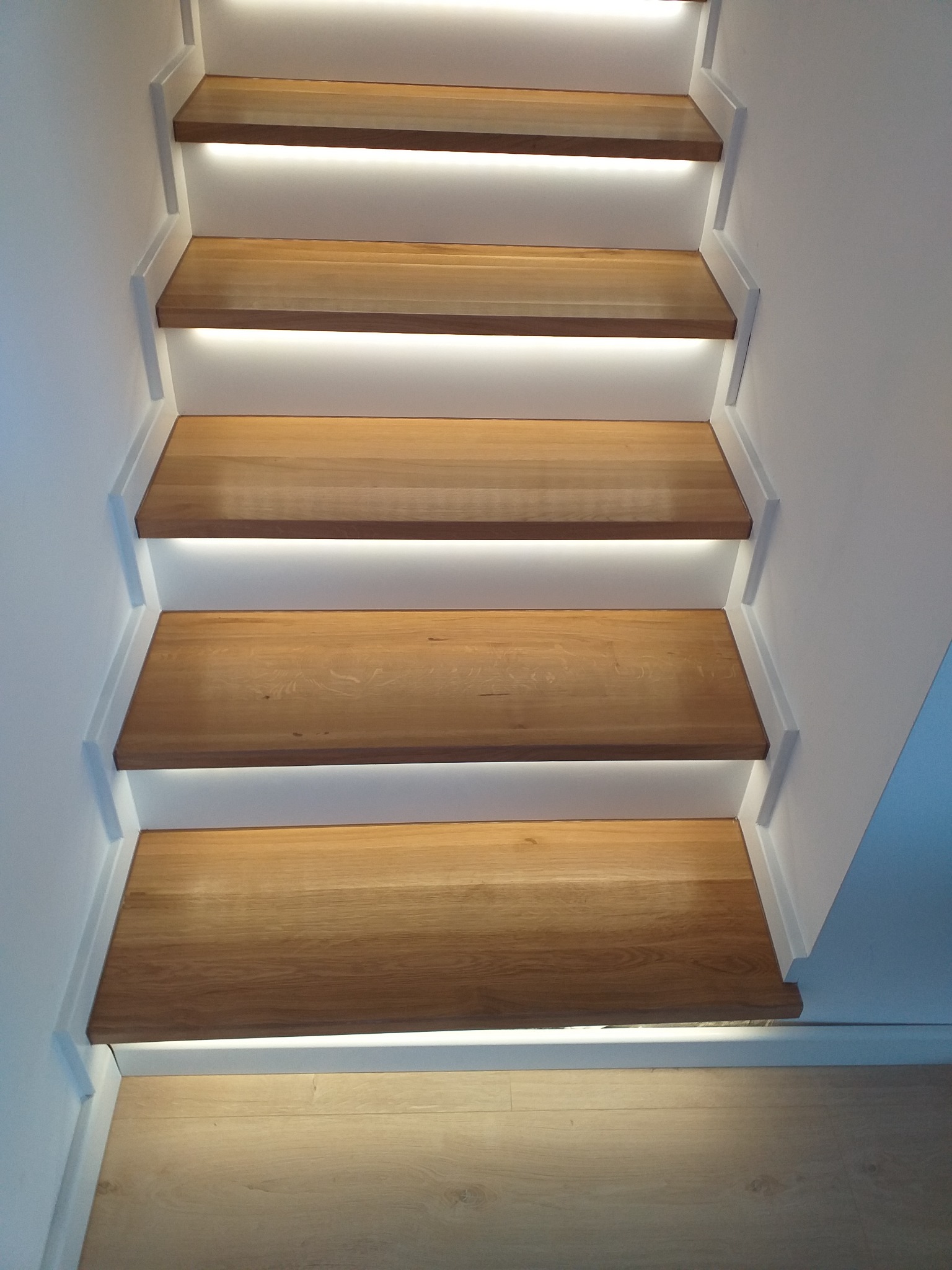 Nowoczesne schody z drewna z podświetleniem LED, widok z dołu na białe ściany i jasne drewniane stopnie.