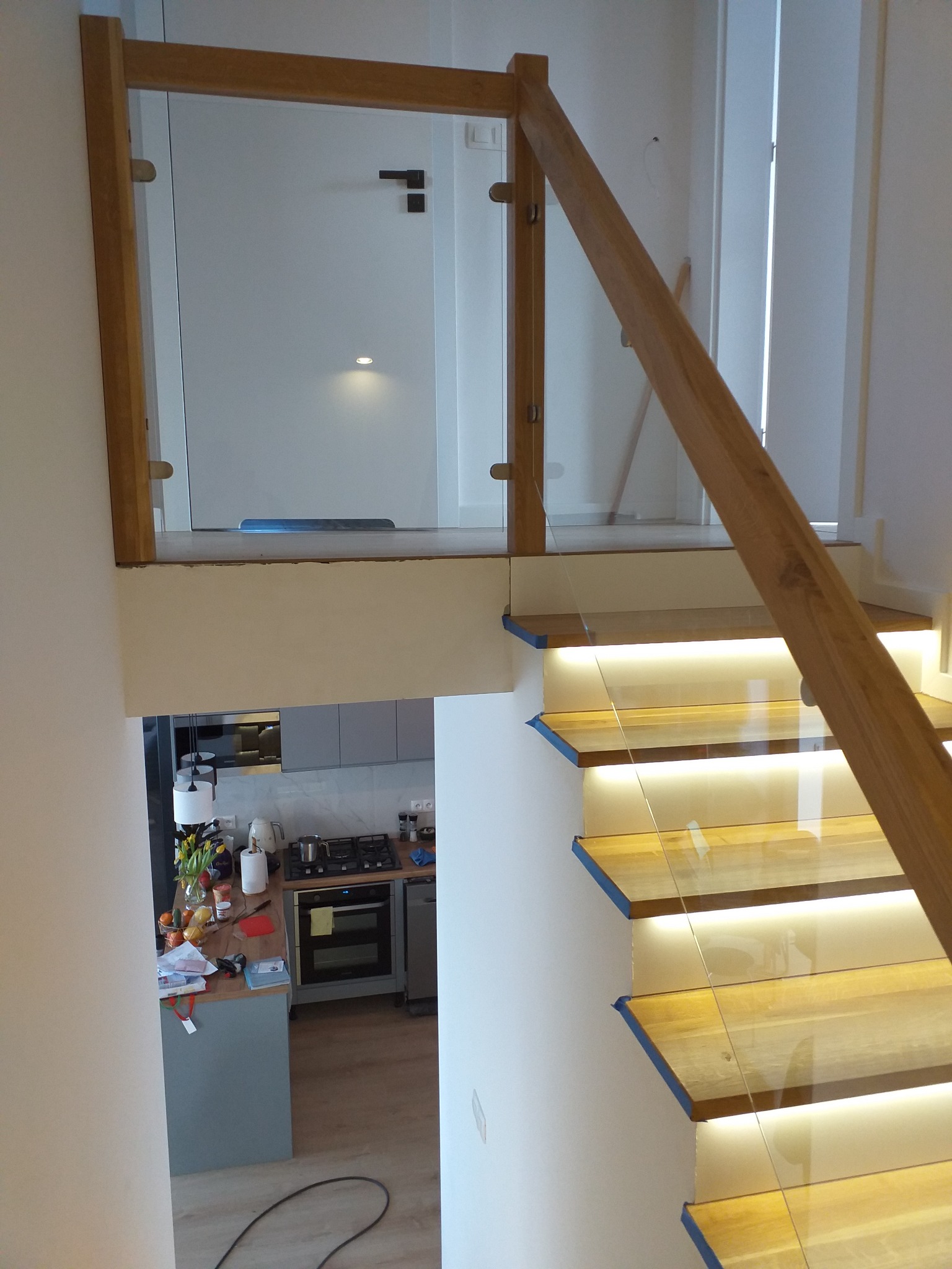 Drewniane schody z podświetleniem LED i szklaną balustradą, widok z góry na fragment kuchni.